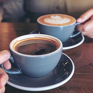 Auf einem Tisch stehen zwei Tassen Kaffee, die von zwei Händen gerade hochgenommen werden