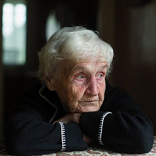 Eine betagte Frau m it kurzen grauen Haaren lehnt auf dem Küchentisch und schaut nachdenklich aus dem Fenster