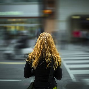 Eine Frau steht vor einer befahrenen Straße. Sie hat offenes Haar. Wir sehen ihren Rücken.