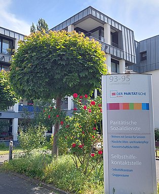 Im Hintergrund: Seniorenwohnanlage. Im Vordergrund ein Baum und eine Infosäule des Paritätischen Bonn und Selbsthilfe-Kontaktstelle