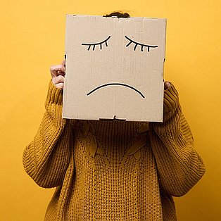 Person trägt Karton pber dem Kopf, auf dem ein trauriges Gesicht gemalt ist