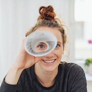 Eine junge Frau blickt uns durch ein zur Rolle gefaltetes Papier an und lächelt
