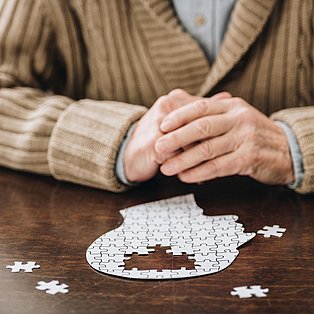 Zu sehen ist eine Person mit einem Puzzle vor sich. Das Puzzle stellt ein menschliches Gehirn dar, in dem einige Teile fehlen.